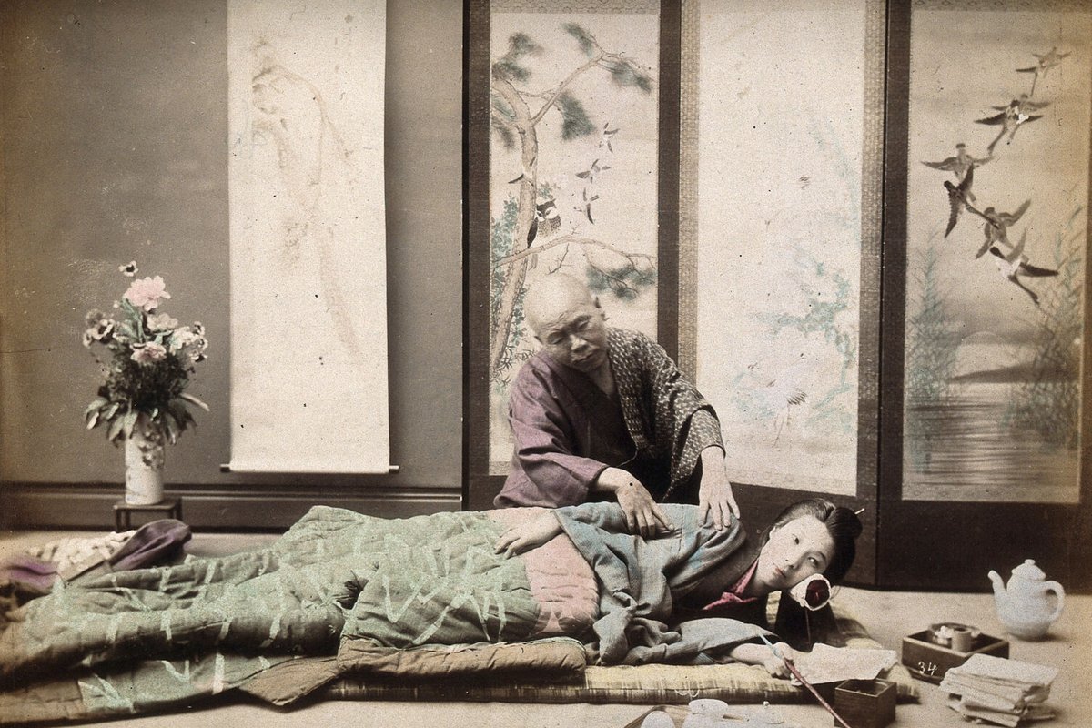Photographie ancienne coloriée : une femme allongée sur un futon, un masseur âgé agenouillé près d'elle, la main sur son dos
