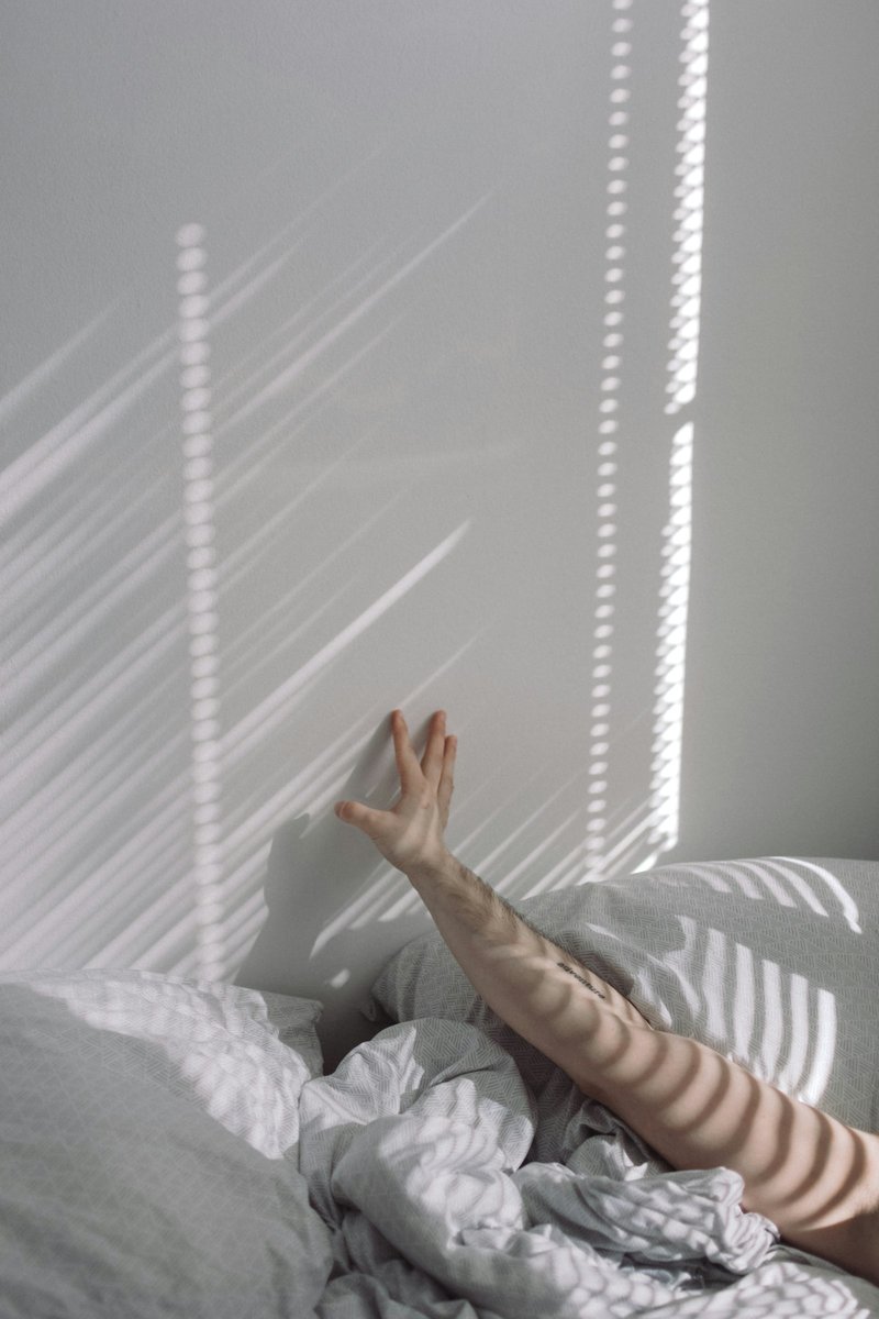 Bras tendu vers le plafond au-dessus d'une couette froissée, strié par les rayons du soleil