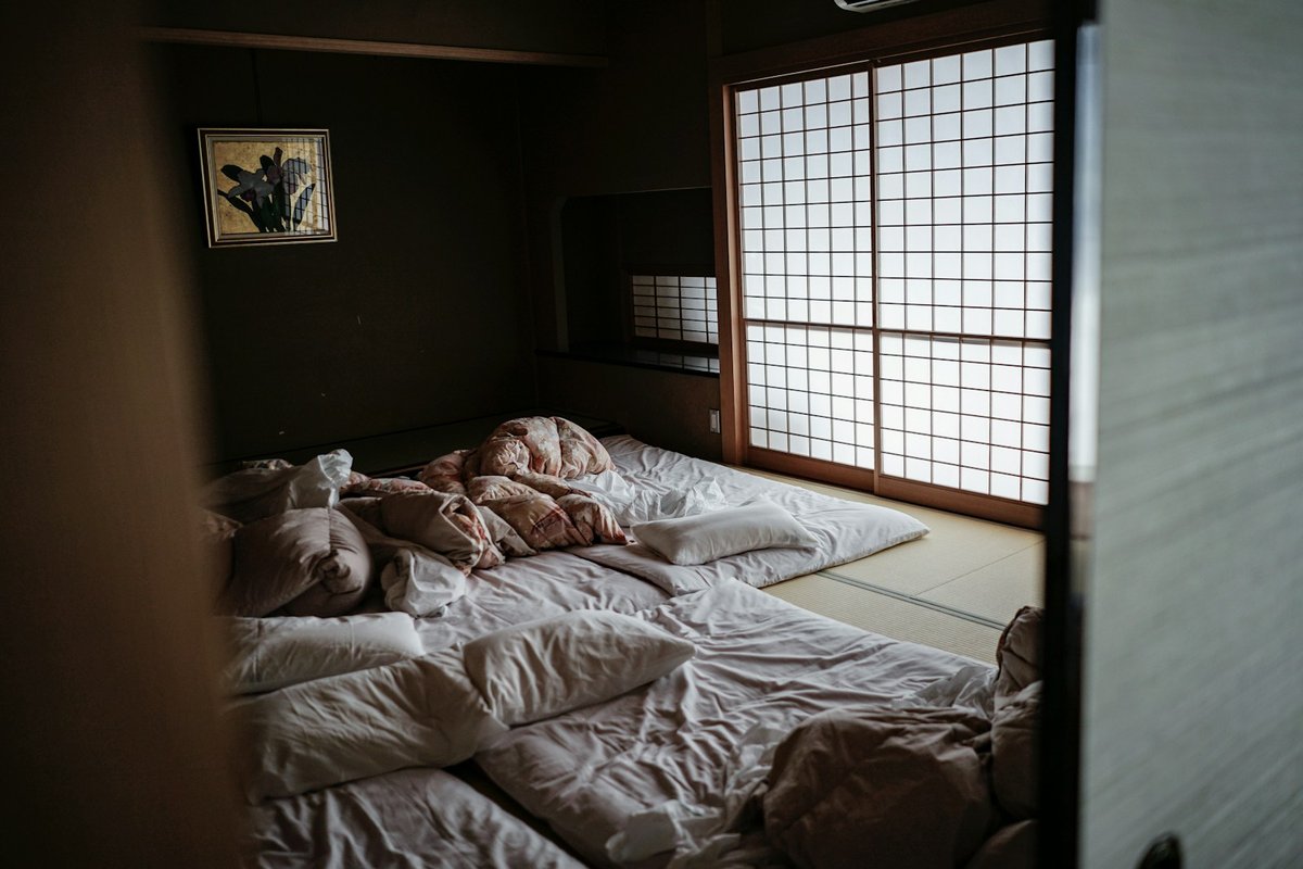 Chambre japonaise dans la pénombre, futons dépliés sur les tatamis près d'une fenêtre à panneaux de papier